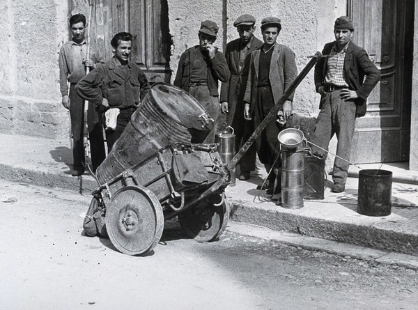 Malaria control equipment, Italy: a large metal barrel in a cart and men on the pavement with more equipment. Photograph, 1940/1950 (?).