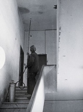 Malaria control equipment, Italy: an apartment building hallway is treated by a man holding a hose attached to a metal canister. Photograph, 1940/1950 (?).