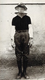 Mosquito headnet and long protective gloves modelled by a man posed in front of a wall. Photograph, 1900/1920.