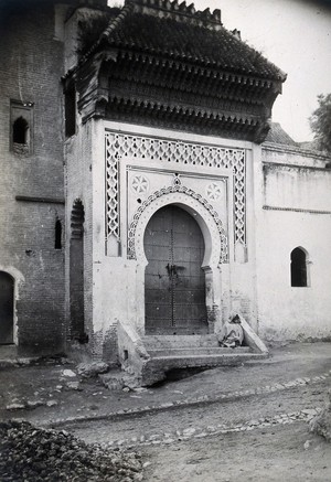 view Ornate doorway of the Medersa (Koranic college) of the Bab Ghissa Mosque, Fez, Morocco; male cloaked figure sitting on the steps. Photograph, ca. 1922.