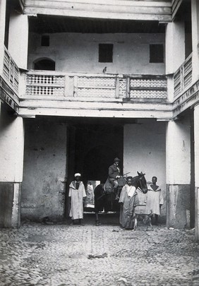 The Martial Director of Hygiene Services (?), on muleback with three standing Moroccan men, outside the medical preventorium (?), before restoration work has begun, Fez, Morocco. Photograph, ca. 1922.