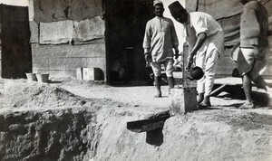 view Soakage pit on a military camp (?) in Egypt (?); an Arab man pours water into the pit's gutter from a kettle, outside wooden huts. Photograph by J.D. Graham, 1914/1918 (?).