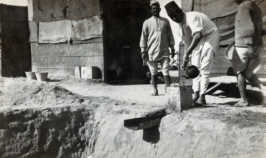Soakage pit on a military camp (?) in Egypt (?); an Arab man pours water into the pit's gutter from a kettle, outside wooden huts. Photograph by J.D. Graham, 1914/1918 (?).