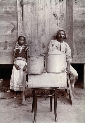 view Milk pails - metal with small taps - with a man and girl in Trinidadian dress, Port of Spain, Trinidad. Photograph, 1900/1920 (?).