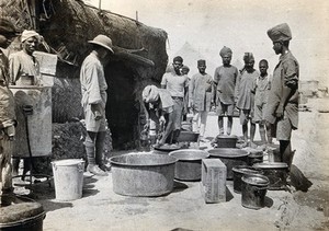 view Basra, Iraq: the Indian General Hospital: milk is served from large metal bowls to waiting men outside the Indian cook house (a straw hut). Photograph, 1914/1918 (?).