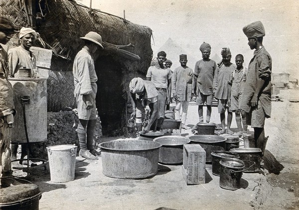 Basra, Iraq: the Indian General Hospital: milk is served from large metal bowls to waiting men outside the Indian cook house (a straw hut). Photograph, 1914/1918 (?).