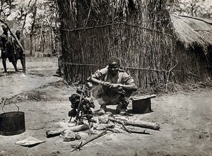 view The Kilwa area (Nanganachi village ?), Tanzania, East Africa: a Tanzanian man spit-roasting meat over an open fire in front of a grass hut. Photograph by Andrew Balfour, ca. 1910 (?).