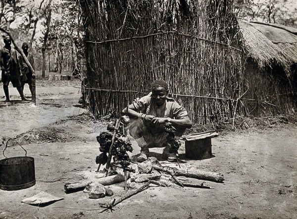 The Kilwa area (Nanganachi village ?), Tanzania, East Africa: a Tanzanian man spit-roasting meat over an open fire in front of a grass hut. Photograph by Andrew Balfour, ca. 1910 (?).