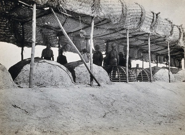 A first world war military camp field bakery: a row of earthen ovens covered by a straw canopy roof, with British and Indian (?) (turbaned) soldiers. Photograph, 1914/1918 (?).