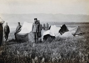 view A First World War military camp (?): a military priest (?), (wearing a black hat with a cross), stands in front of tents in a field with mountains in the background; several other soldiers nearby. Photograph, 1914/1918 (?).