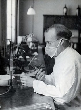 Yellow fever: a scientist wearing a face mask works at a laboratory bench with bunsen burners and microscopes. Photograph, 1910/1930 (?).