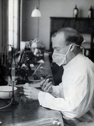 view Yellow fever: a scientist wearing a face mask works at a laboratory bench with bunsen burners and microscopes. Photograph, 1910/1930 (?).