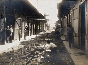 view Panama (?): a man retrieves his hat from a ditch running down the middle of a street lined with wooden buildings. Photograph, ca. 1910.