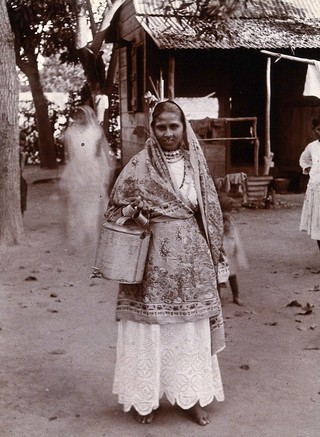 A Trinidadian woman in traditional costume carrying a metal milk pail with a tap, Port of Spain, Trinidad. Photograph, 1905/1915.