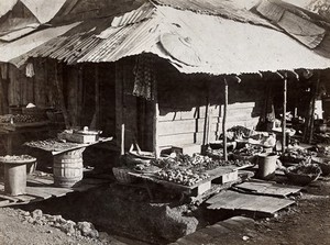 view Shops in Freetown, Sierra Leone: wooden huts with food displayed outside, over open drains. Photograph, ca. 1911.