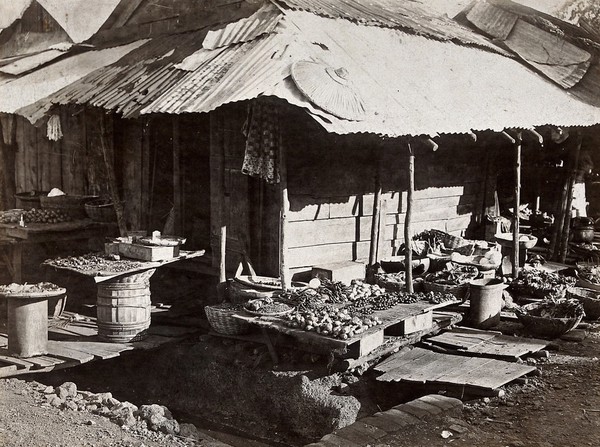 Shops in Freetown, Sierra Leone: wooden huts with food displayed outside, over open drains. Photograph, ca. 1911.