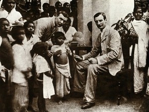 view Inoculation against cholera: two men in suits prepare to inoculate a child, surrounded by a crowd of (Indian ?) parents and children. Photograph, 1880/1900.