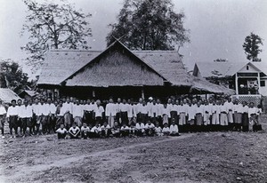 view Chiengmai Leper Asylum, Thailand: a group of patients outside the temporary grass-roofed chapel; the asylum overseer's house to one side. Photograph, 1921.