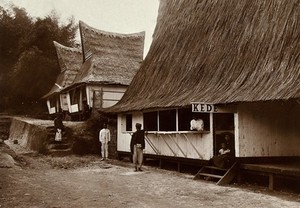 view Boarding houses for visitors to a local asylum for people with leprosy, next to the village shop; the buildings have tall, pointed thatched rooves. Photograph, 1890/1910.