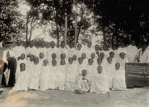 view Leper settlement, Fianarantsoa, Madagascar: a group portrait of patients in white robes, with an elderly woman (missionary ?); one patient holds up a blackboard bearing a bible verse (in Malagasy ?). Photograph, 1890/1910.