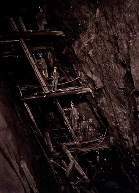 Dolcoath tin mine, Camborne, Cornwall: miners in a mine shaft, standing on wooden platforms connected by ladders. Photograph by J. C. Burrow, 1890/1910.