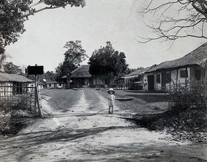 view The Kalar-Azar Commission Camp field laboratory, Golaghat, Assam, India: a small, white boy in a large hat stands at the camp entrance; grass-roofed buildings with Indian men in the background. Photograph, 1910/1930 (?).