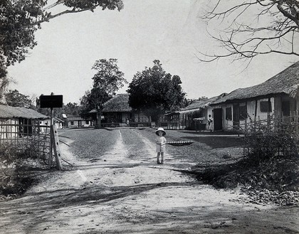 The Kalar-Azar Commission Camp field laboratory, Golaghat, Assam, India: a small, white boy in a large hat stands at the camp entrance; grass-roofed buildings with Indian men in the background. Photograph, 1910/1930 (?).