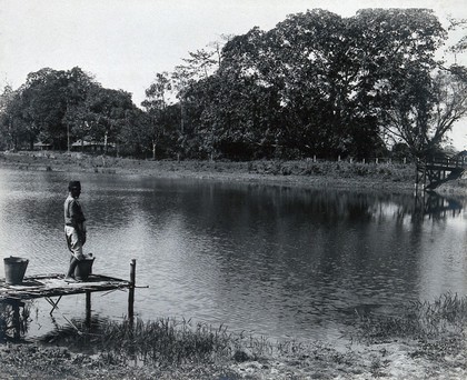 A reservoir, India: an Indian man stands with two buckets on the jetty. Photograph, 1910/1920 (?).