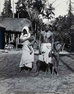 view Assam, India: an Indian man and woman, wearing simple robes, with their two small children, outside a grass-roofed house. Photograph, 1900/1920 (?).