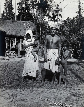 Assam, India: an Indian man and woman, wearing simple robes, with their two small children, outside a grass-roofed house. Photograph, 1900/1920 (?).