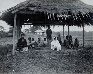 view Assam, India: a kala-azar out-patient centre: a small group of men, women and children sit beneath a grass-roofed shelter with some metal bowls (for heating water for sterilisation ?). Photograph, 1900/1920 (?).