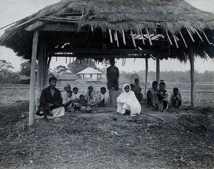 Assam, India: a kala-azar out-patient centre: a small group of men, women and children sit beneath a grass-roofed shelter with some metal bowls (for heating water for sterilisation ?). Photograph, 1900/1920 (?).