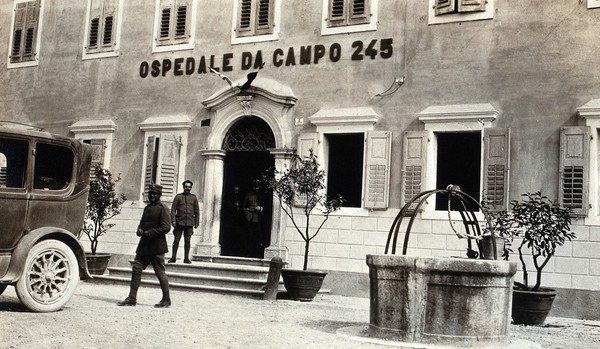World War One: an Italian field hospital with soldiers standing at the entrance and a well outside. Photograph, 1917.