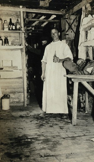 view World War One: [Bougaincourt?], France: a dressing station in a dug-out: a medic wearing a white gown with a patient on a makeshift bed. Photograph, 1916.