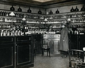 view The Worshipful Society of Apothecaries' pharmacy, Water Lane, Blackfriars, London: a woman is served by the pharmacist, who stands behind a high polished wood counter, in front of shelves holding glass pharmacy jars. Photograph, ca. 1935.
