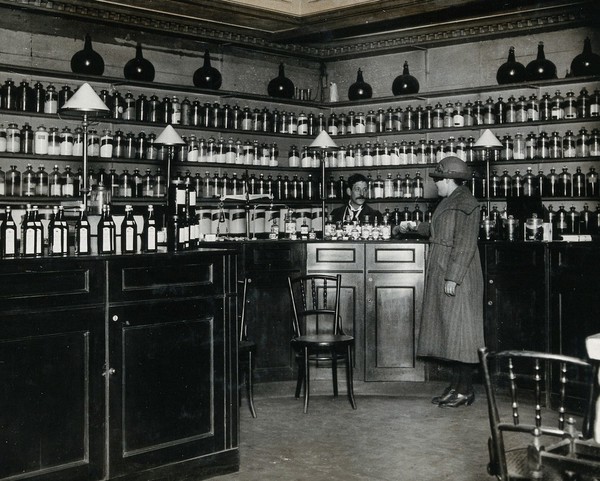 The Worshipful Society of Apothecaries' pharmacy, Water Lane, Blackfriars, London: a woman is served by the pharmacist, who stands behind a high polished wood counter, in front of shelves holding glass pharmacy jars. Photograph, ca. 1935.
