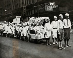 view Pennsylvania, U.S.A.: a dental hygiene parade: children parade wearing caps shaped liked teeth, bearing banners reading "We brush our teeth each day 100%" and "Progress first". Photograph, 1910/1920.