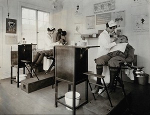 view Dental clinic, U.S.A.: dental hygienists examine two boys seated in wooden dentist chairs; the room is decorated with dental hygiene awareness posters. Photograph, ca. 1920.