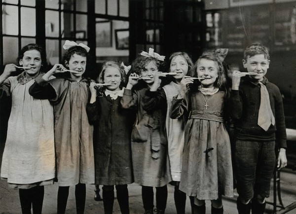 Tooth-brush drill at school, England: children pose with tooth-brushes in mouth in a school building. Photograph, ca. 1920.