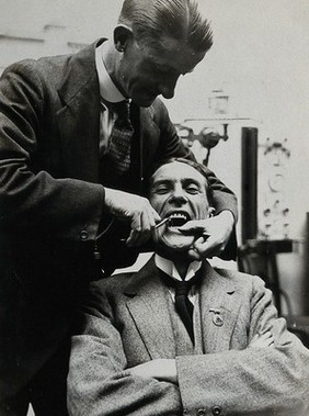 Dental extraction: a dentist prepares to use a metal implement to extract the tooth of a grinning man. Photograph, ca. 1921.