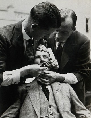 Dental extraction: two men hold a man's head still and mouth open to demonstrate out-dated dental extraction techniques. Photograph, ca. 1921.