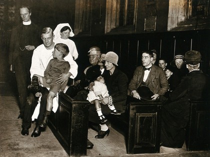 A bonesetter, A.E. Kennard, at work in St Katherine Cree church, London: a small boy with a bound leg sits on his lap while patients wait in the pews; a nurse and a vicar look on. Photograph, ca. 1925.