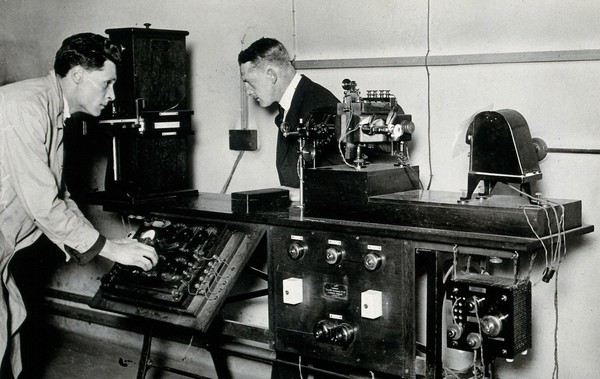 An electrocardiograph at the National Hospital for Diseases of the Heart, London: one man adjusts the dials while another looks on. Photograph, ca. 1922.