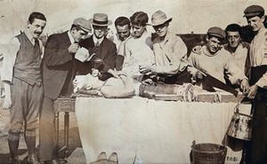 view Staging of a leg amputation: the 'patient' lies on a table surrounded by men: one man poses with a saw, two men pretend to administer pain relief applying a teapot and a funnel to the patient's mouth. Photograph, 1905.