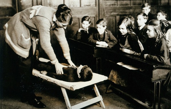 A First Aid lesson in a school classroom: the teacher demonstrates with a small boy lying on a stretcher, watched by children seated in rows at wooden desks. Photograph, ca. 1920.