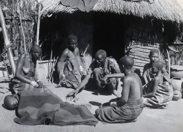 Africa: a medicine man sits with a circle of people including a sick child under a blanket. Photograph by N.J. Van Warmelo, ca. 1935.