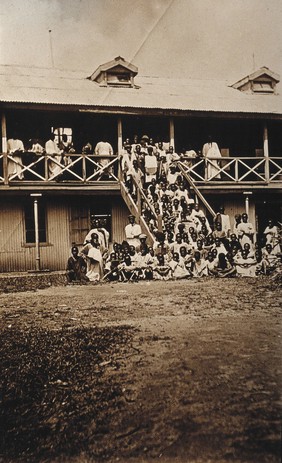 Accra, Ghana: African people waiting to be inoculated on the porch and steps of a (hospital?) building. Photograph, 1910/1920.
