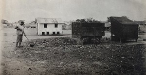 view Lagos, Nigeria: latrine buildings and a man seen sweeping rubbish with a broom. Photograph, 1910/1920.