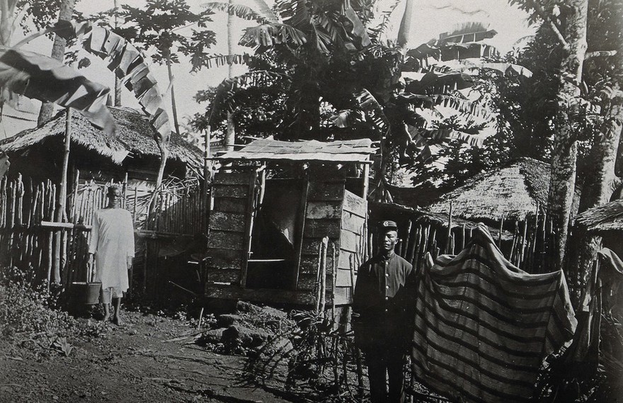 Sierra Leone: wooden cesspool huts, with a uniformed man and a man holding a bucket shown standing in the foreground. Photograph, 1910/1920.