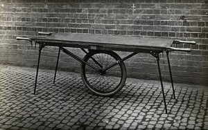 view Closes' mono-wheel stretcher, in open position: side view, showing the stretcher resting on four fold-out legs, with one large central wheel in contact with the ground. Photograph, 1915/1918.